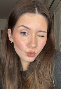 Nuori nainen, jolla on pitkät ruskeat hiukset, iskee silmää ja pusertaa huulet, hänellä on korvissa renkaskorvakorut ja septum-koru, kuvattu sisätiloissa neutraalin taustan edessä.