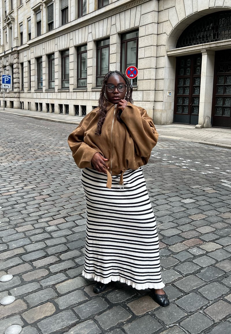 Jeune personne avec des cheveux tressés et des lunettes, portant une veste marron et une jupe rayée noir et blanc, debout dans une rue pavée.