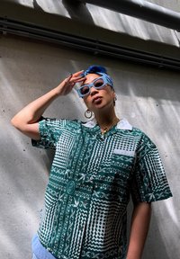 Camisa de manga corta de color verde con un diseño estampado y patrones geométricos blancos, con cuello. Combinada con gafas de sol azules y un pañuelo azul.