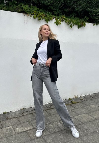 Blazer negro, camiseta blanca y jeans de pierna ancha gris claro. Zapatillas blancas. De pie contra una pared blanca con vegetación arriba.