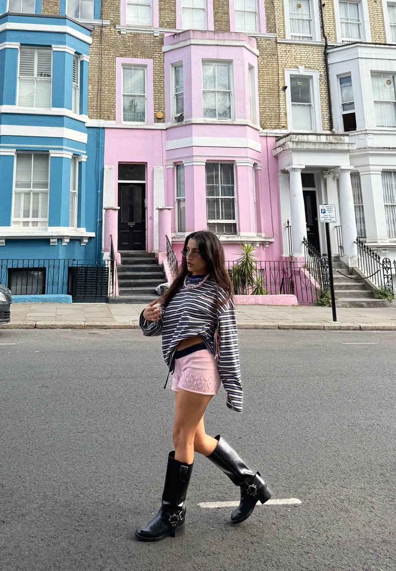 Femme marchant dans la rue devant des rangées de maisons colorées bleu, rose et blanc, portant un haut rayé, un short rose et des bottes noires.
