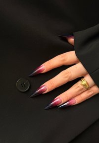 Long, pointed nails featuring a gradient from pink to dark purple are showcased against a black fabric background, accompanied by a black button and a gold ring.