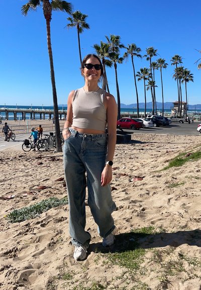 Top cropped beige claro, jeans azules holgados y zapatillas blancas. El fondo presenta palmeras, playa y un muelle bajo un cielo azul despejado.