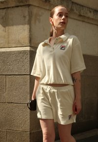 Polo shirt beige, à texture douce, avec des manches courtes et un col, assorti à un short. Présente un petit logo sur la poitrine.