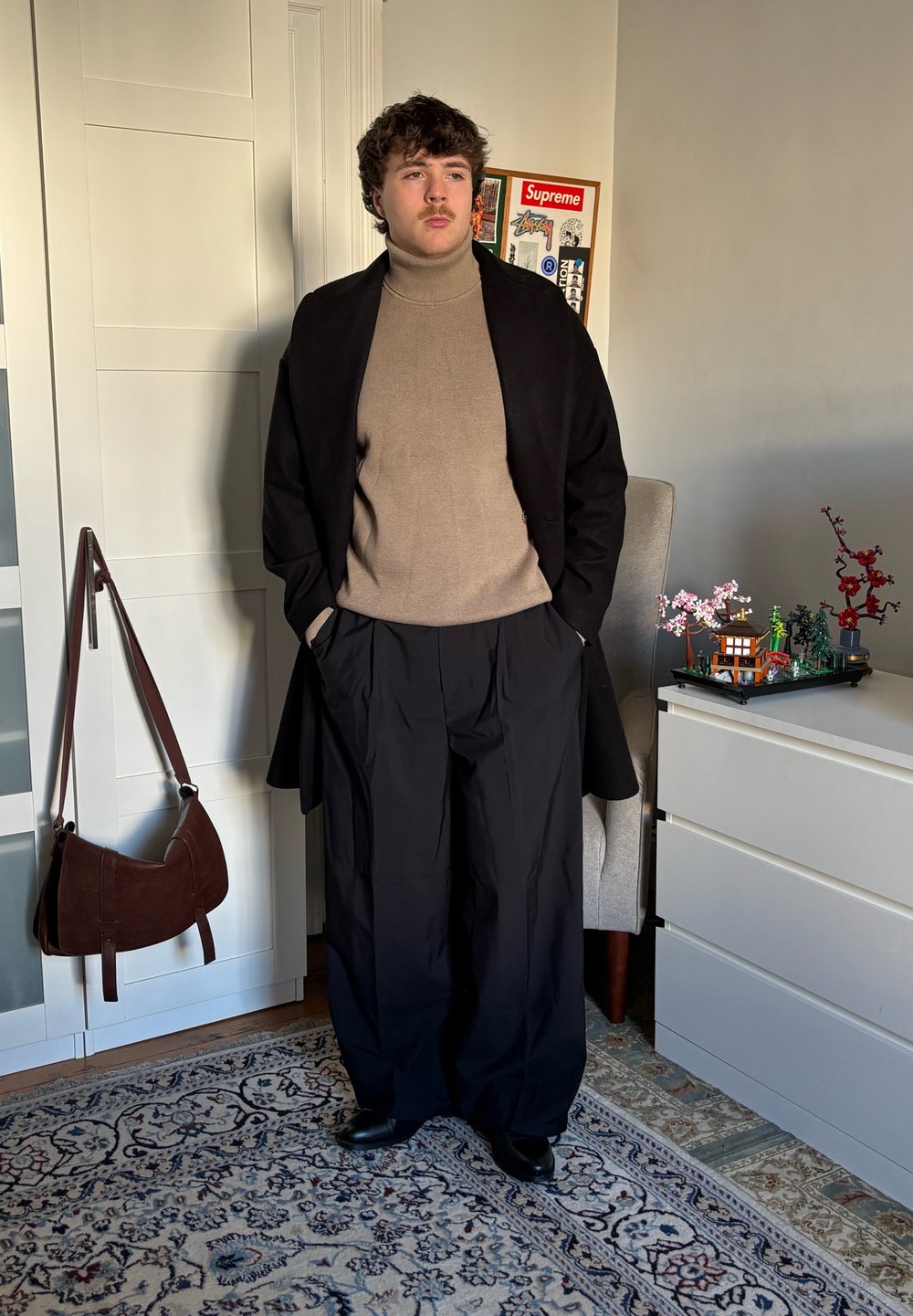 Sort frakke over beige rullekrave, løse sorte bukser og sorte sko. Brun skuldertaske hængende i nærheden. Neutralt farvet indretning i baggrunden.