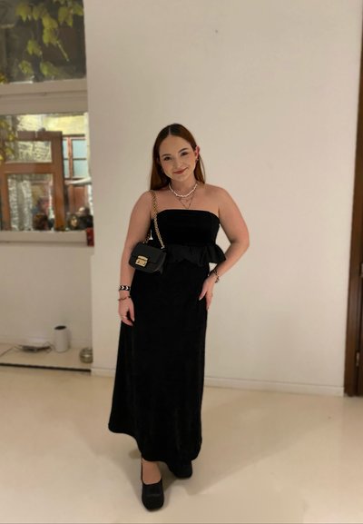 Vestido maxi negro sin tirantes con una cintura con volantes, combinado con un pequeño bolso negro y accesorios. Textura suave, color sólido, silueta elegante.