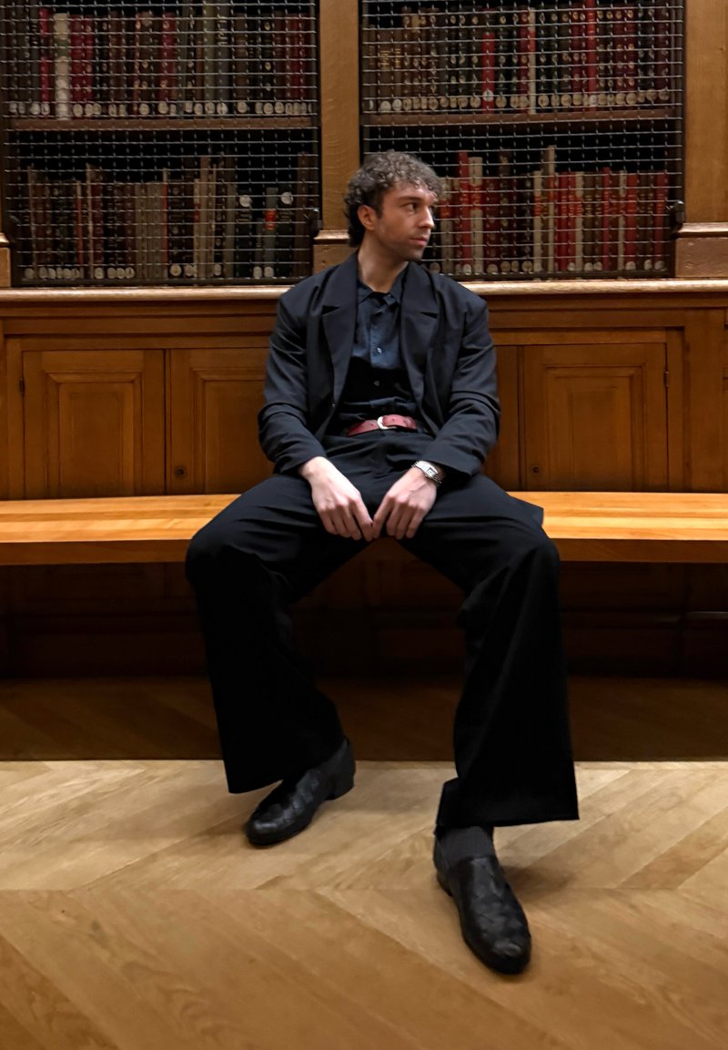 Homme en costume noir assis sur un banc en bois, regardant de côté, avec des armoires en bois et des livres derrière lui.