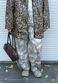 Veste imperméable à motif léopard, pantalon à motifs gris et blancs, sac à main marron, baskets grises, avec des feuilles tombées au sol.