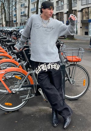 Suéter gris de canalé con corte holgado y mangas a rayas, combinado con jeans negros que presentan letras llamativas y un detalle de cadena. Botas negras de punta.