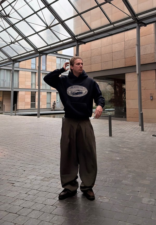 Homme portant un grand sweat à capuche sombre et un pantalon large, debout sous une verrière dans une cour urbaine, ajustant ses cheveux d'une main.