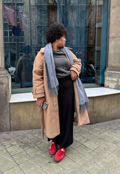 Abrigo beige, suéter gris, pantalones negros, bufanda azul claro con flecos y zapatillas rojas. De pie contra una ventana de vidrio.