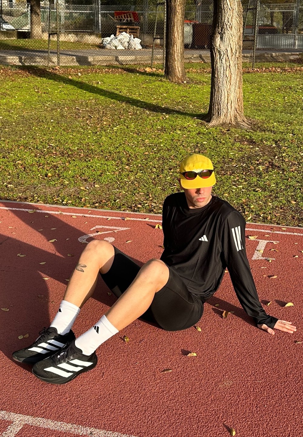 Camiseta deportiva negra de manga larga con rayas blancas, pantalones cortos negros, gorra amarilla y gafas de sol, sentado en una pista de atletismo roja con césped.