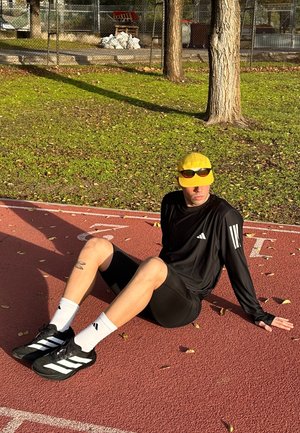 Camiseta deportiva negra de manga larga con rayas blancas, pantalones cortos negros, gorra amarilla y gafas de sol, sentado en una pista de atletismo roja con césped.