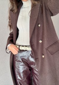 Άτομο που φοράει λευκό ριμπ ζιβάγκο, σκούρο καφέ παλτό με κουμπιά, λαμπερό σκούρο δερμάτινο παντελόνι, λευ κή ζώνη και χέρι στην τσέπη, μπροστά σε απλό τοίχο.