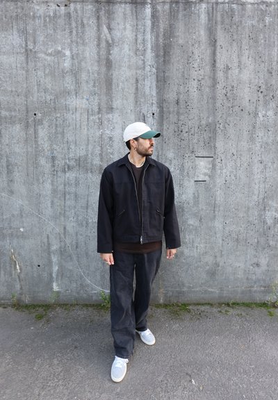 Chaqueta negra con cremallera sobre una camisa marrón, pantalones oscuros y zapatillas grises. Sombrero color crema con visera verde. Pared de hormigón y suelo pavimentado al fondo.
