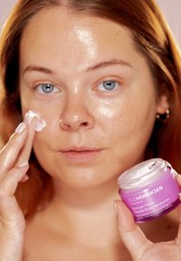 Lila burk med Ole Henriksen fuktkräm hålls i ena handen, medan den andra handen applicerar kräm på ett glansigt ansikte. Krämig konsistens, genomskinligt lock.