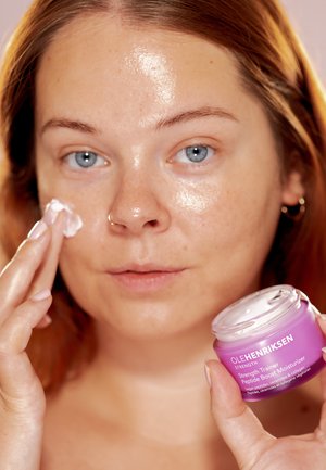 Barattolo viola di crema idratante Ole Henriksen tenuto in una mano, mentre l'altra applica la crema su un viso luminoso. Tessuto cremoso, coperchio trasparente.