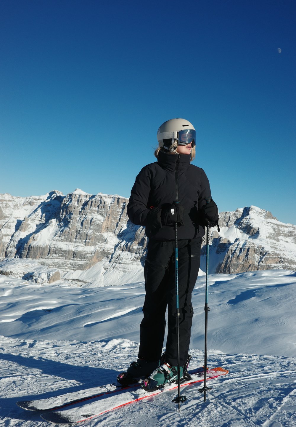 Équipement de ski avec une veste et un pantalon isolants noirs, un casque beige et des lunettes de ski, debout sur la neige avec des bâtons de ski devant un paysage montagneux.