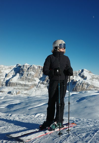 Equipo de esquí con chaqueta y pantalones negros aislantes, un casco beige y gafas de esquí, de pie sobre la nieve con bastones de esquí frente a un paisaje montañoso.
