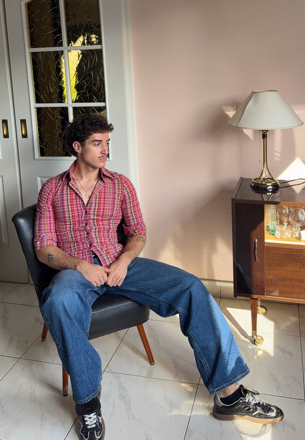 Hombre con cabello rizado y bigote que lleva una camisa de cuadros roja y vaqueros azules sentado en una silla negra en una habitación iluminada por el sol con pared rosa y armario de madera.