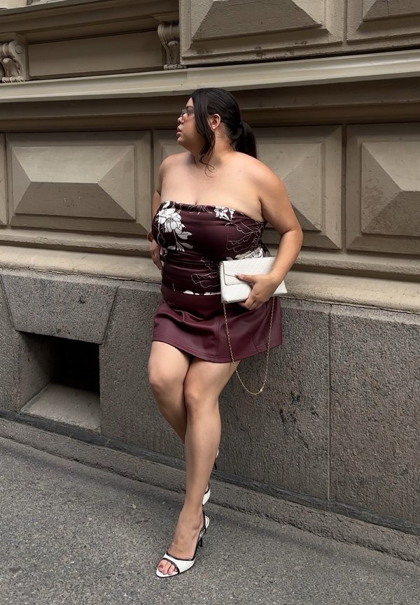 Femme portant des lunettes, un haut fleuri sans bretelles et une jupe bordeaux, tenant une pochette blanche, appuyée contre un mur en pierre dans la rue.