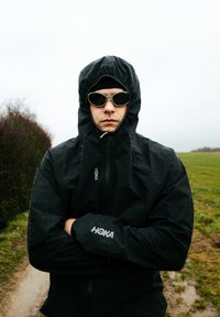 Personne portant une veste de pluie à capuche noire et des lunettes de soleil, debout sur un chemin rural mouillé avec de l'herbe et des buissons à proximité.