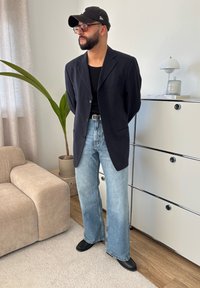 Blazer riscado preto sobre uma t-shirt preta, jeans flare azul claro e sapatos pretos. De pé ao lado de uma planta e mobiliário moderno.
