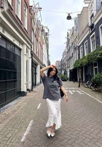 Grijze oversized t-shirt, witte kant rok met lagen, puntige zilveren schoenen, smal bakstenen pad, omringd door gebouwen en groen.