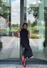 Μαύρη αμάνικη μπλούζα με ψηλό λαιμό και εφαρμοστή σιλουέτα σε συνδυασμό με μακριά μαύρη φούστα. Οι εντυπωσιακές κόκκινες γόβες με ψηλό τακούνι αναδεικνύουν το σύνολο.