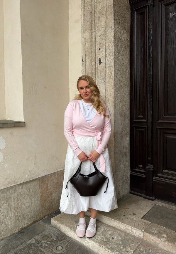 Tânără cu cardigan roz și rochie albă care ține o geantă neagră, stând pe trepte de piatră lângă o ușă de lemn și un perete bej.