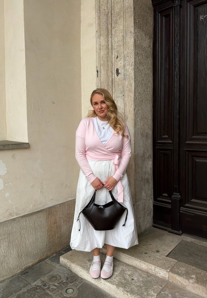 Junge Frau in einem rosa Cardigan und weißem Kleid, die eine schwarze Handtasche hält und auf Steinstufen bei einer Holztür und einer beigefarbenen Wand steht.
