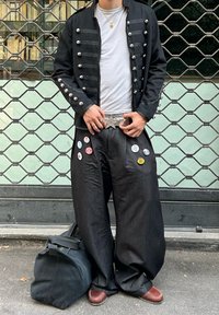 Veste noire style militaire avec boutons argentés, portée sur une chemise blanche. Pantalon noir ample avec badges colorés en forme de boutons. Sac sombre posé au sol.