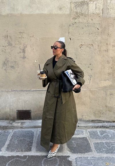 Mujer con un abrigo trench largo de color oliva, gafas de sol negras y tacones con manchas, sosteniendo un café y una revista, de pie sobre un pavimento de piedra junto a una pared desgastada.
