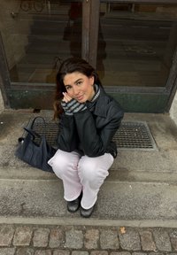 Veste en cuir noir, écharpe grise rayée, pantalon rose clair, bottes noires. Assis sur des marches en pierre avec un sac à main sombre à proximité.