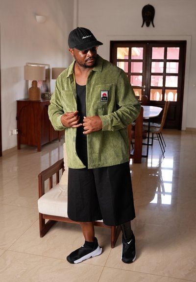 Camisa de pana verde con un logo en el pecho, camiseta sin mangas negra, pantalones cortos negros sueltos, zapatillas negras y una gorra negra. Entorno interior neutral.