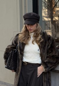 Γυναίκα που φοράει καφέ παλτό από γούνα, λευκό ζιβάγκο με πλεκτό μοτίβο καλωδίου, μαύρο παντελόνι, μαύρο καπέλο και κουβαλάει μαύρη πλεκτή τσάντα σε εξωτερικό χώρο.