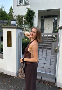 Brązowy top na ramiączkach z koralików i ciemne szerokie spodnie, stojąca blisko szarej metalowej bramy przed białym domem otoczonym zielenią.