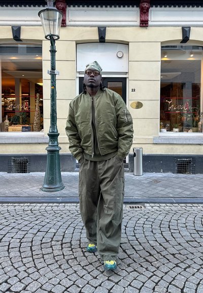 Chaqueta bomber de color verde oliva con puños acanalados, pantalones holgados a juego y zapatillas con acentos azules y amarillos; de pie en una calle de adoquines.