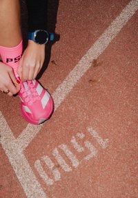 Élénk rózsaszín sportcipő fehér csíkokkal, rózsaszín fűzővel bekötve. Kék okosórát visel a csuklóján, piros futópálya felület előtt.