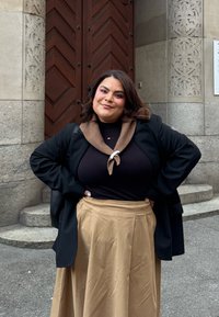 Lang beige nederdel, sort top og sort blazer med brune kravedetaljer. Stående foran udsmykkede trædøre og stenarkitektur.