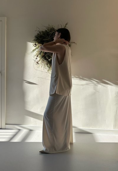 Top sin mangas de un beige claro con aberturas laterales, combinado con una falda maxi fluida de tela suave y un brillo sutil, frente a una pared neutra.