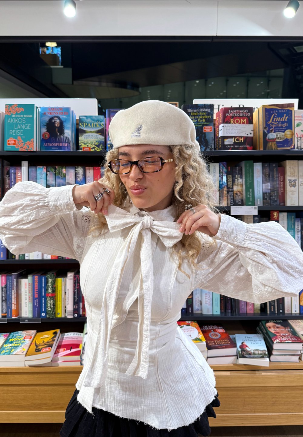 Blusa bianca strutturata con ampie maniche e fiocco al colletto, abbinata a un basco bianco. Scaffali di libri con varie copertine colorate sullo sfondo.