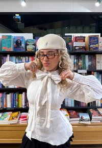 Blusa bianca strutturata con ampie maniche e fiocco al colletto, abbinata a un basco bianco. Scaffali di libri con varie copertine colorate sullo sfondo.