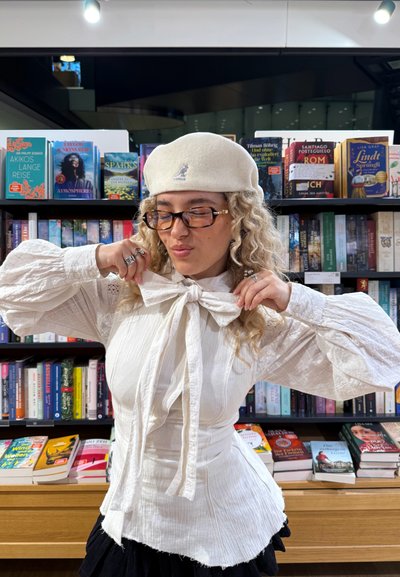 Blusa blanca texturizada con mangas anchas y un lazo en el cuello, combinada con una boina blanca. Estanterías con libros de diversas portadas coloridas al fondo.