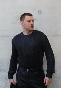 Musta palmikkoneulepusero, jossa on pyöreä kaula-aukko, teksturoitu kuviointi ja pitkät hihat, yhdistettynä mustiin nahkahousuihin. Taustaväri on harmaa.