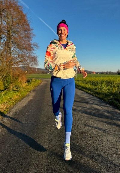 Chaqueta ligera y con estampado sobre una camiseta atlética azul y leggings; zapatillas de running con detalles en blanco y verde claro; fondo de cielo despejado.