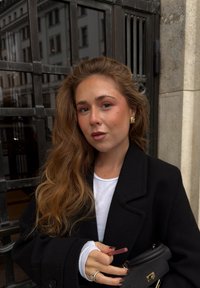 Svart ullkåpe over en hvit skjorte, holder et rosa leppeprodukt. Langt, bølget brunt hår, gulløreringer og svart håndveske med gullbeslag.