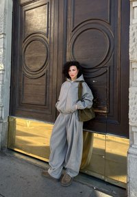 Kvinde med krøllet hår iført rummeligt gråt tøj og brune sko læner sig op ad store udsmykkede trædøre med gylden metalbase.