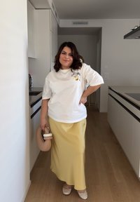 Fehér, rövidujjú póló logóval, egy lenge, sárga selyem maxi szoknyával párosítva. Bézs táska a kézben és semleges balerina cipők. Modern konyhát háttérként.