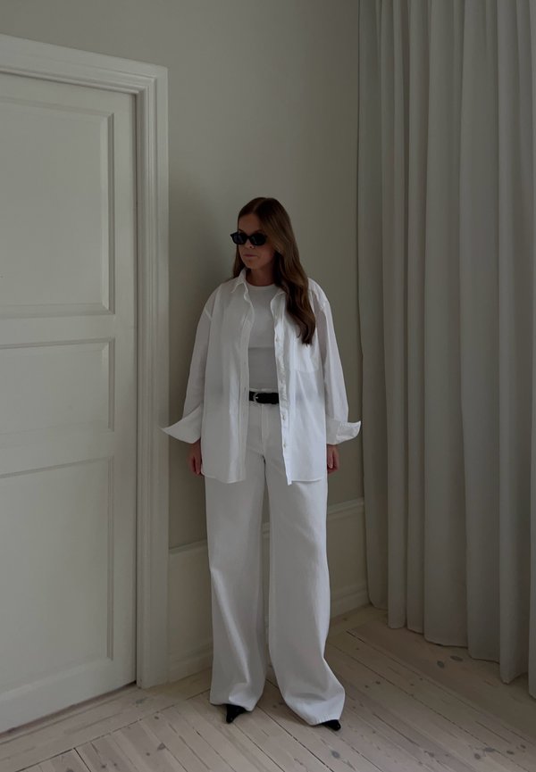 Femme portant une chemise blanche oversize, un pantalon blanc à jambes larges, une ceinture noire et des lunettes de soleil noires, debout à l'intérieur près d'une porte fermée et de rideaux.